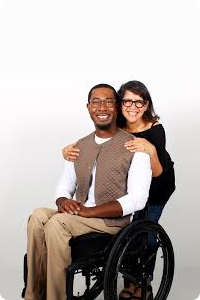 A man in a wheelchair wearing an IZ adaptive outfit of seamless back elastic waist beige chino trousers and brown waistcoat, with a smiling woman standing behind him.